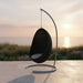 Black rattan outdoor hanging egg chair with a white cushion on a metal stand overlooking the ocean.
