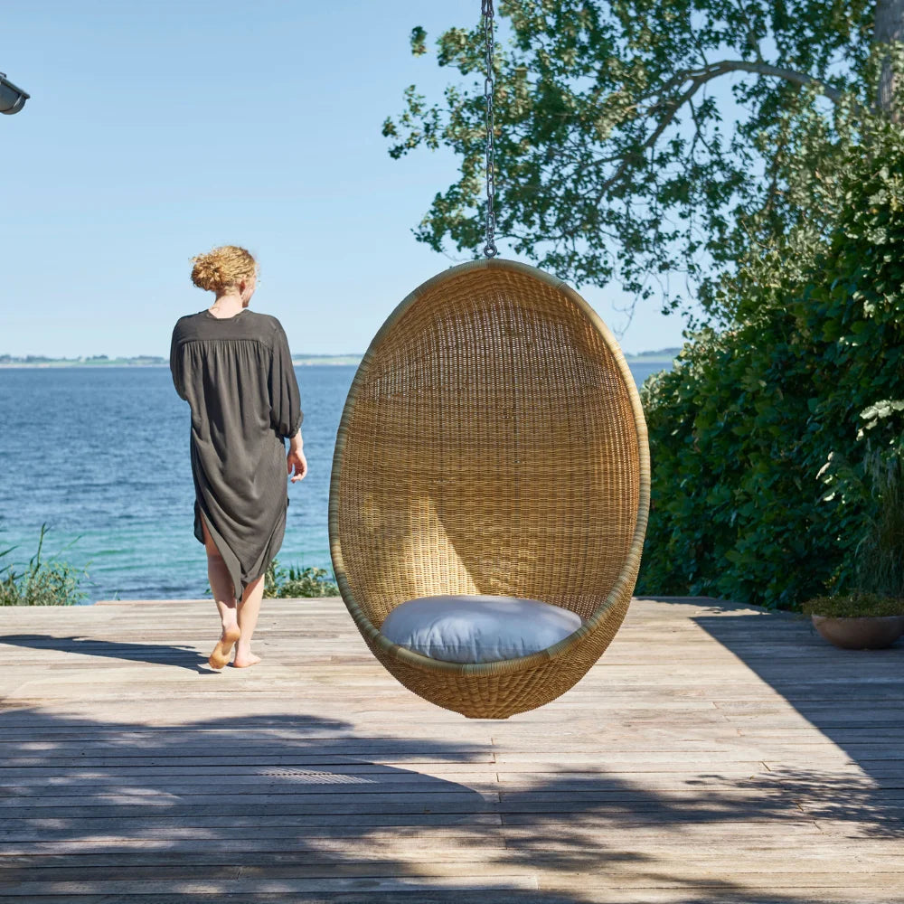 Natural rattan outdoor hanging egg chair with a white cushion beside a woman on a seaside deck.