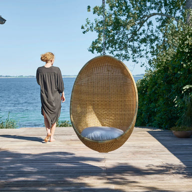 Natural rattan outdoor hanging egg chair with a white cushion beside a woman on a seaside deck.