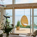 Natural rattan outdoor hanging egg chair with a white cushion in a bright room overlooking the ocean.