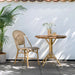 Round teak outdoor café table beside a matching natural rattan dining side chair on a patio.