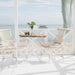 Round teak outdoor café table with white base between matching high-back lounge chairs on a seaside patio.