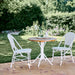 Round teak outdoor café table with white base beside matching white rattan dining side chairs on a garden patio.