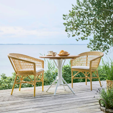Round teak outdoor café table with white base matching natural rattan dining armchairs on a seaside deck.