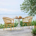 Round teak outdoor café table with white base matching natural rattan dining armchairs on a seaside deck.