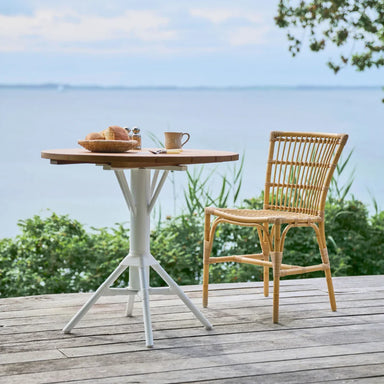 Round teak outdoor café table with white base matching natural rattan dining side chair on a seaside deck.
