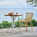 Round teak outdoor café table with white base matching natural rattan dining side chair on a seaside deck.