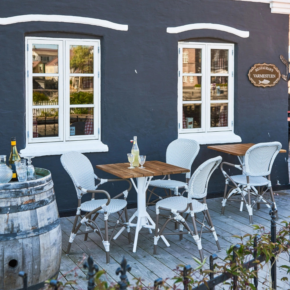 Square teak outdoor café table with a white pedestal base beside matching white rattan dining armchairs.