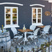 Square teak outdoor café table with a white pedestal base beside matching white rattan dining armchairs.