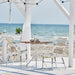 Square teak outdoor café table with a white pedestal base beside matching white rattan dining armchairs.