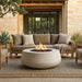 Outdoor patio furniture set with a round concrete fire pit in the center, a beige sectional sofa, matching armchairs, and a woven rug in front of a stone patio building.