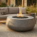 A round concrete fire pit table with a glowing flame and black fire rocks, set in an outdoor patio area next to a gray sectional sofa.