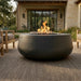 A round dark gray concrete fire pit table with active orange flames, set on a textured outdoor rug in front of a modern stone house.