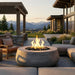 A round concrete fire pit with a glowing fire in the center, set on a modern outdoor patio with lounge chairs, a wooden pergola, and a mountain landscape in the background.