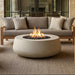 A round concrete fire pit table with a glowing flame and black stones, placed in front of a beige outdoor sofa on a woven rug.