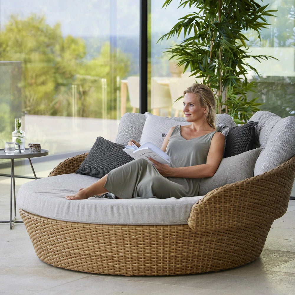 A woman relaxes in a large round woven wicker daybed with thick light gray cushions, set in a modern patio lounge area.