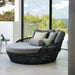 A round wicker daybed with a gray cushion and multiple accent pillows, placed indoors near large glass windows overlooking greenery.