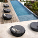 Four round outdoor lounge chairs with dark woven frames and gray cushions are arranged along a wooden deck beside a blue swimming pool.