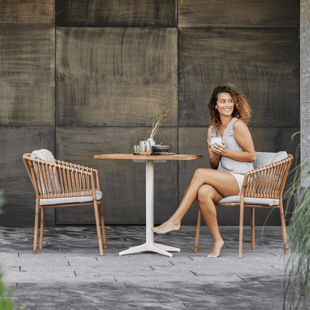 Boxhill's Ocean Outdoor Dining Armchair lifestyle image at patio with teak table and a woman sitting holding a cup of coffee