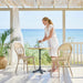 Natural rattan outdoor dining chair beside a square teak café table with a woman on a seaside patio.