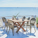 Gray rattan outdoor dining side chairs beside a rectangular teak dining table on a seaside patio.

