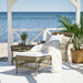 Gray rattan outdoor chaise lounge with white cushions and a matching side table on a seaside patio.