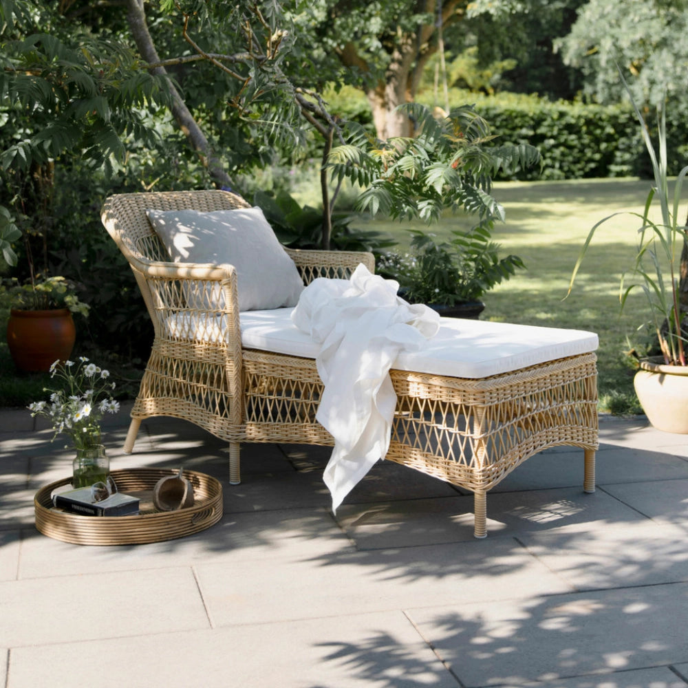 Natural rattan outdoor chaise lounge with white cushions beside a round serving tray on a garden patio.