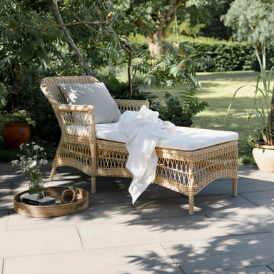 Natural rattan outdoor chaise lounge with white cushions beside a round serving tray on a garden patio.