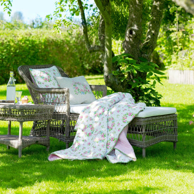 Gray rattan outdoor chaise lounge with white cushions beside a matching side table in a garden.