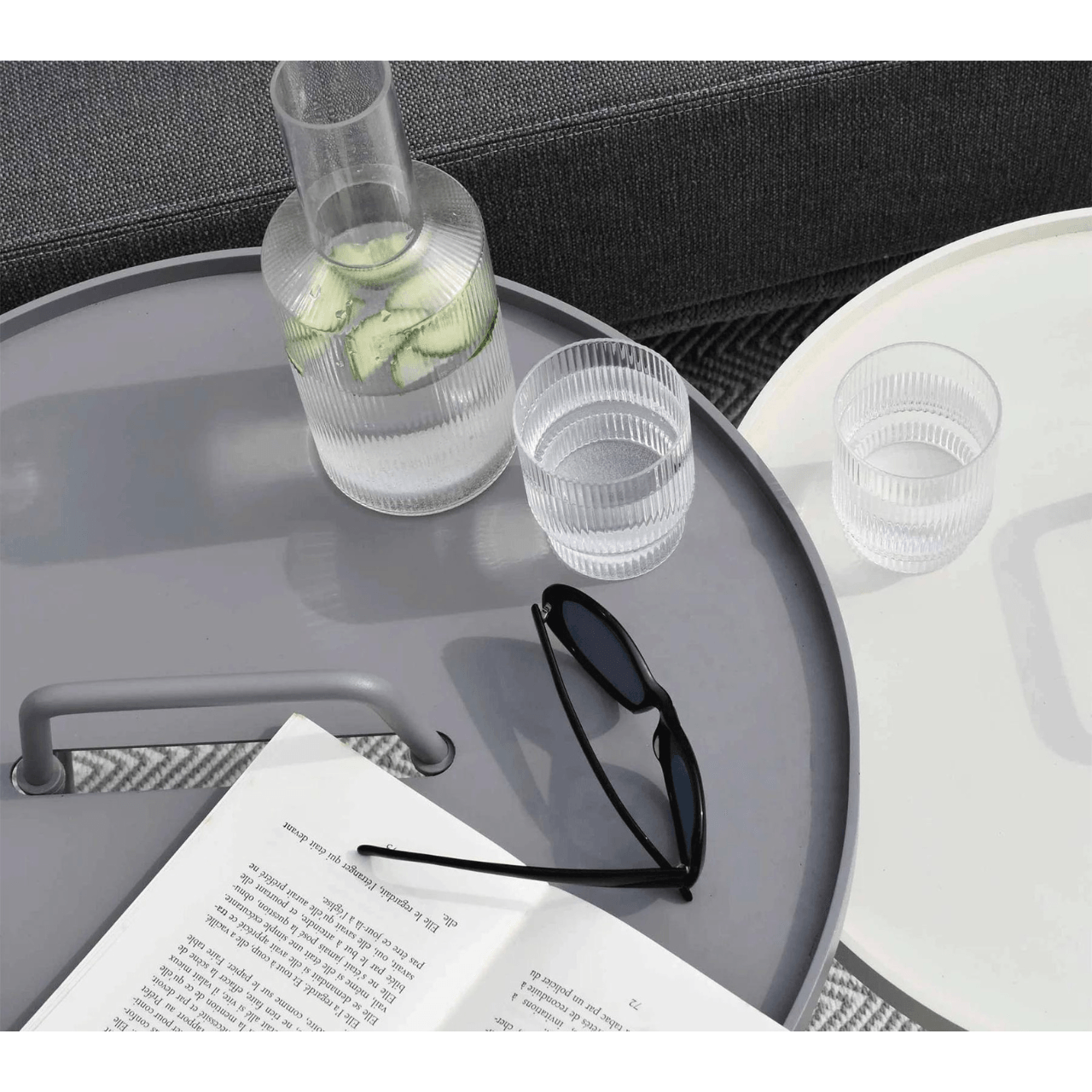 Boxhill's On-The-Move light grey and white outdoor round side table with a glass of water, bottle container, sunglass and a book on it