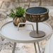 Small round aluminum side table with a woven lamp and potted plant on a stone surface