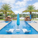 A luxury outdoor swimming pool with bright blue water, two white lounge chairs, and a closed light blue umbrella in the foreground. The pool is bordered by beige stone tiles, flanked by two tall palm trees, rock features with waterfalls, and a lake with houses in the distance under a blue sky with clouds.