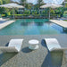 Modern outdoor poolside setting with two white lounge chairs and a small round side table on a stone deck, with large umbrellas, palm trees, and a villa in the background.