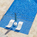 A top-down view of a swimming pool with bright blue water, beige stone deck flooring, two white lounge chairs, and a closed teal-blue patio umbrella on a small round table.