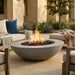 A round concrete fire pit bowl with black river rocks and a visible flame, placed in an outdoor patio setting with lounge chairs, plants, and a building facade in the background.