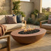 A outdoor concrete oval terracotta-colored outdoor fire pit table with black river rocks and visible flames, placed on a patio between modern lounge chairs.