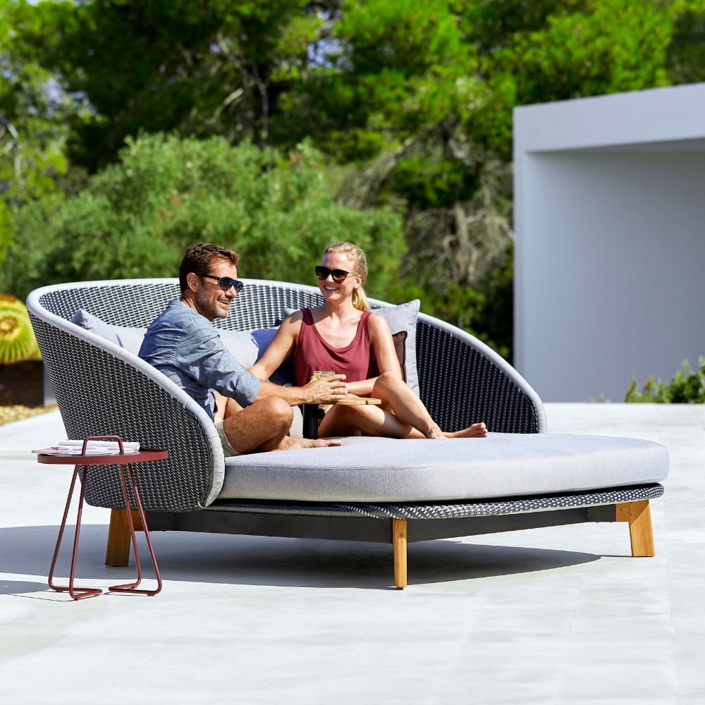 Boxhill's Peacock grey outdoor daybed with man and wowan sitting on it and maroon outdoor round side table at the side