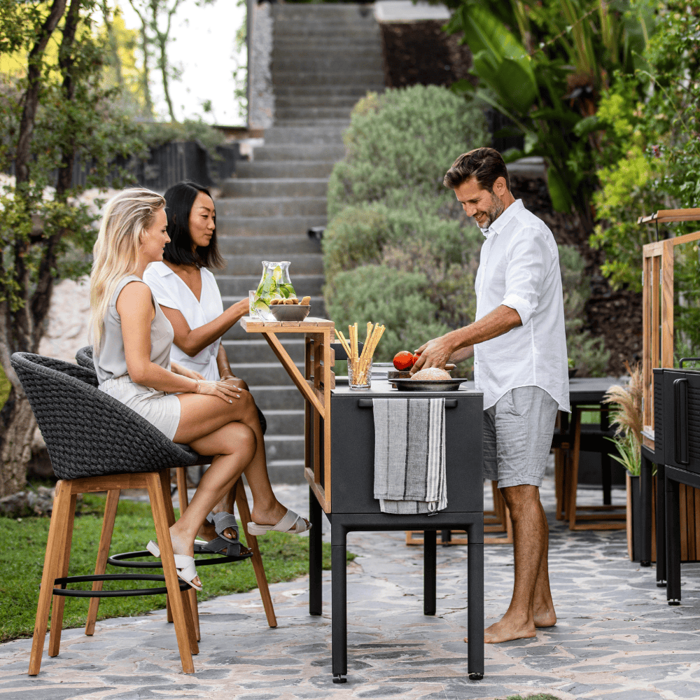 Boxhill's Peacock dark grey outdoor bar chair with teak legs with 2 woman sitting on it and man standing infront of kitchen shelves