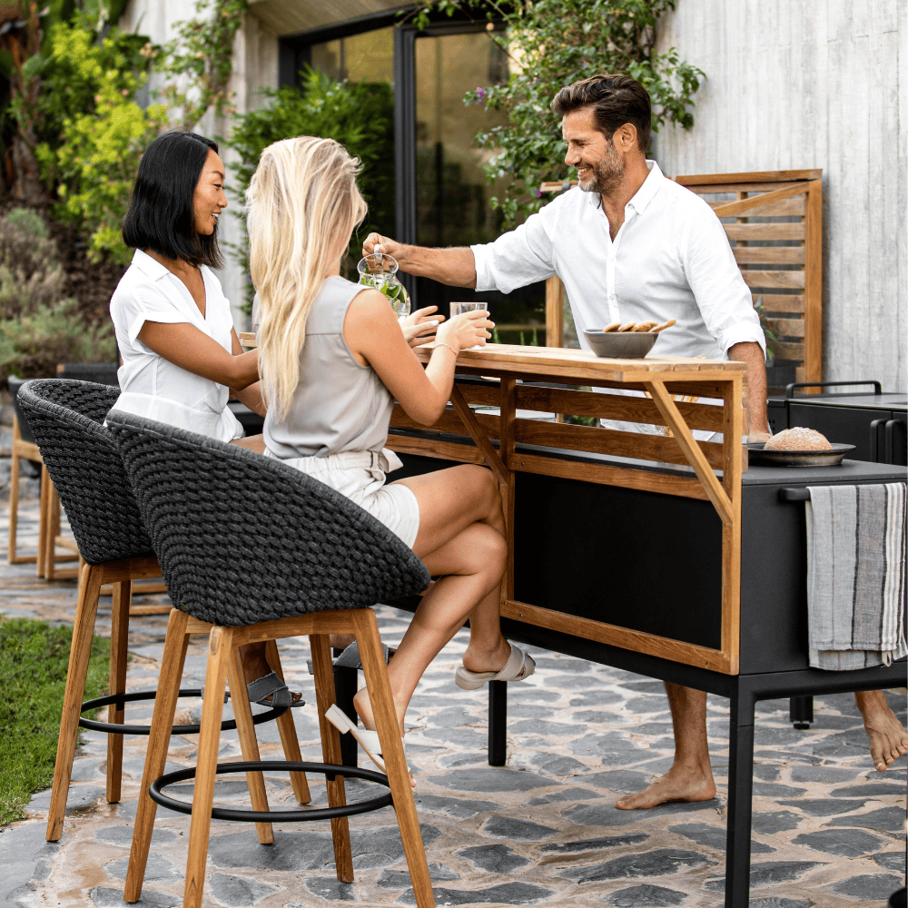 Boxhill's Peacock dark grey outdoor bar chair with teak legs with outdoor kitchen shelves and 3 people having a good time