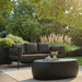 Patio seating with black pebble concrete outdoor coffee table on light beige rug, dark gray sofa, surrounded by greenery