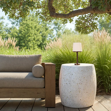 Outdoor seating area with a wooden sofa,  round white concrete side table, small lamp, and lush greenery under a tree
