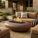 An outdoor patio scene with an oval brown fire pit table in the foreground, flames burning inside, and a tan wicker sofa with cushions and pillows in the background. A house exterior and landscaping are visible.