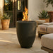 A dark charcoal-colored outdoor fire pit planter with a glowing orange flame, set on a stone patio in front of a modern house.