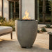 Outdoor tall cylinder-shaped concrete gray fire pit on a patio with a building and plants in the background