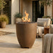 Outdoor patio with a tall cylinder-shaped concrete brown fire pit, chairs, and potted plants.