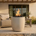 A tall cylindrical outdoor fire pit with a textured light beige stone-like finish, filled with black river rocks and lit with a bright orange and yellow flame, set on a patio in front of a beige house with a sofa and side table.