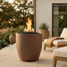 Outdoor patio with a tall cylinder-shaped outdoor concrete brown fire pit, chairs, and potted plants.