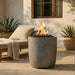 Outdoor setting with a tall cylinder-shaped outdoor concrete gray fire pit table, lounge chair, and potted plants.