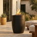 Black tall cylindrical outdoor conrete fire pit with hidden propane enclosure door with flames on a patio with furniture and plants.