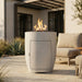 A modern cylindrical outdoor fire pit with a light gray concrete finish and a visible flame, placed on a patio next to a beige cushioned chair in front of a house.